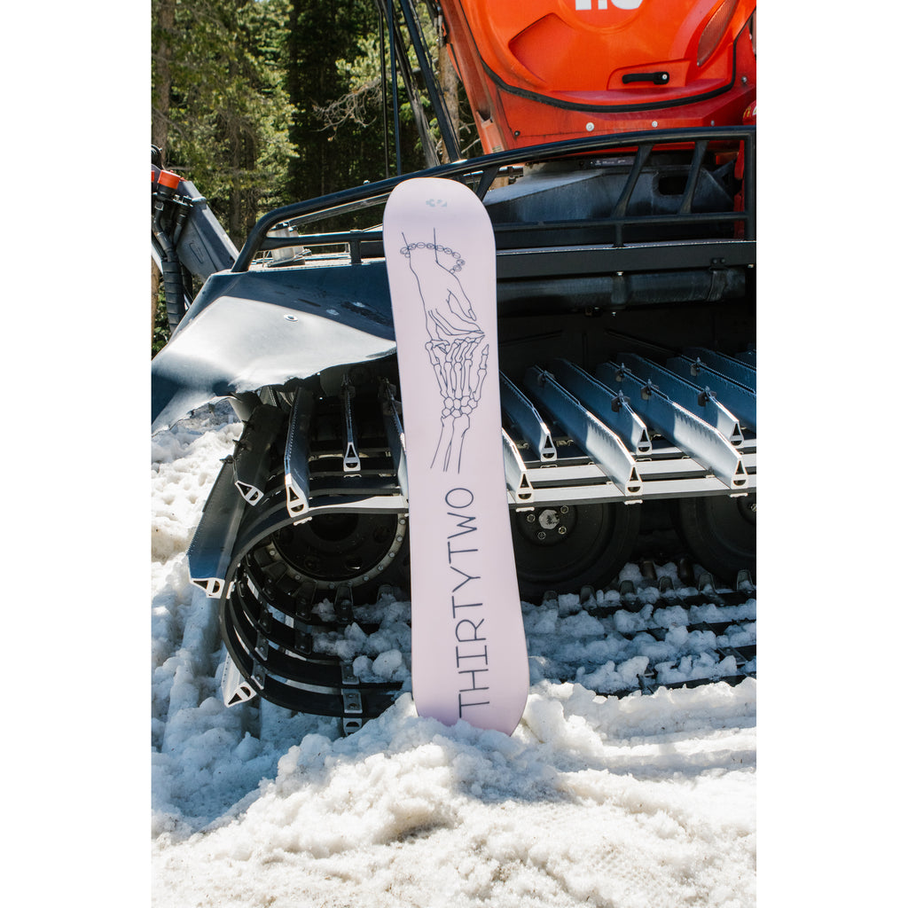 Women’s Crucible snowboard with pink skeleton hand base graphic beside a snowmobile