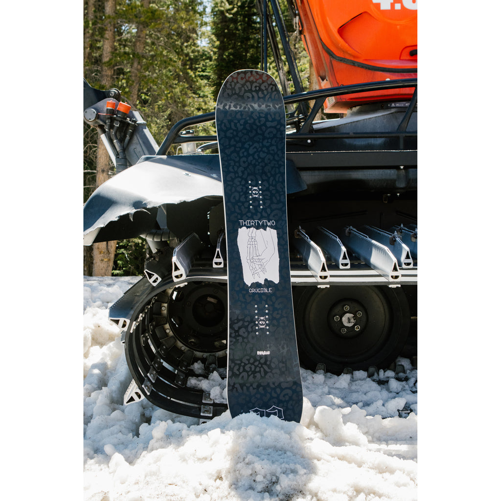 Women’s Crucible snowboard with black skeleton hand topsheet beside a snowmobile